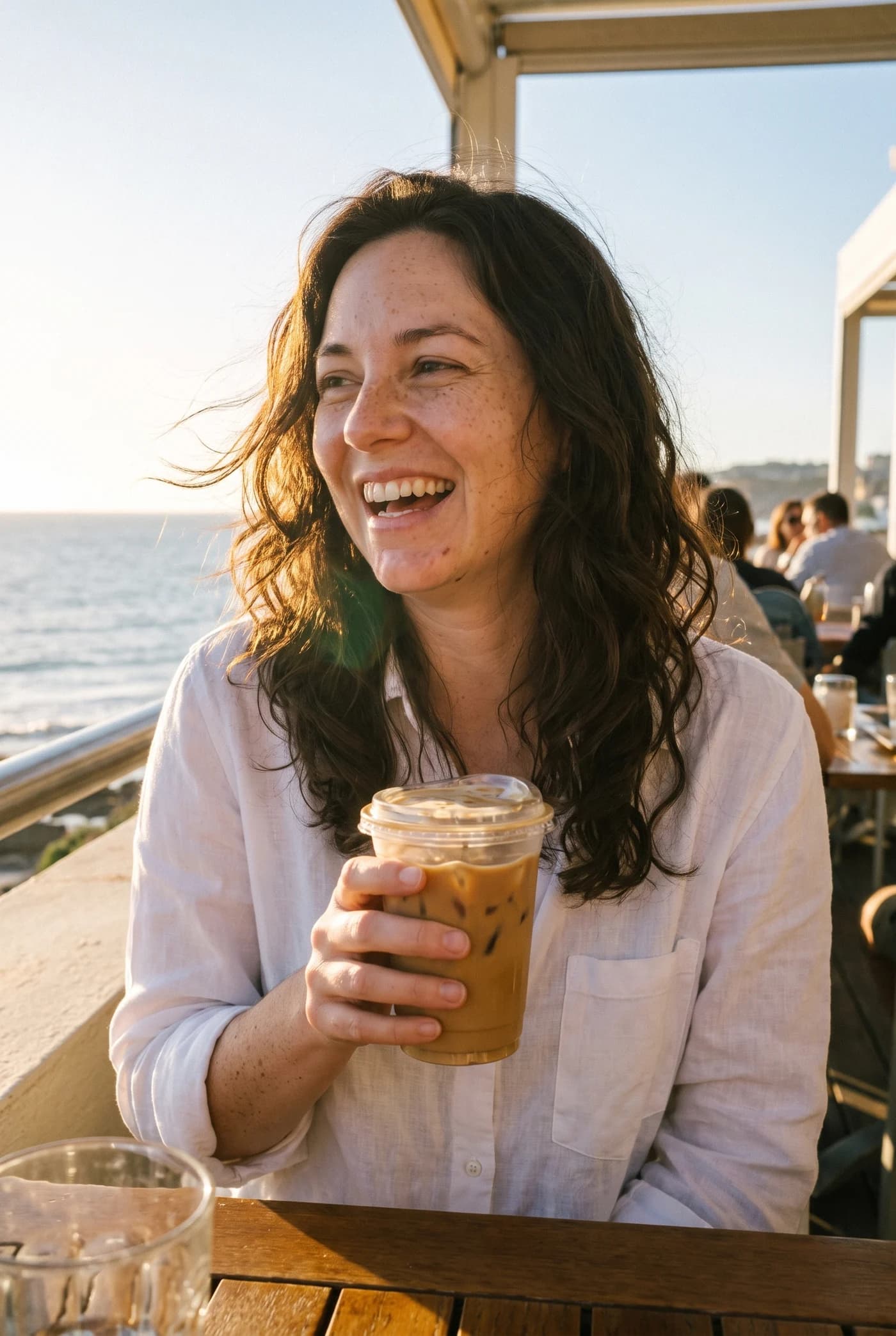 The same woman laughing with an iced coffee on a sunny seaside terrace.