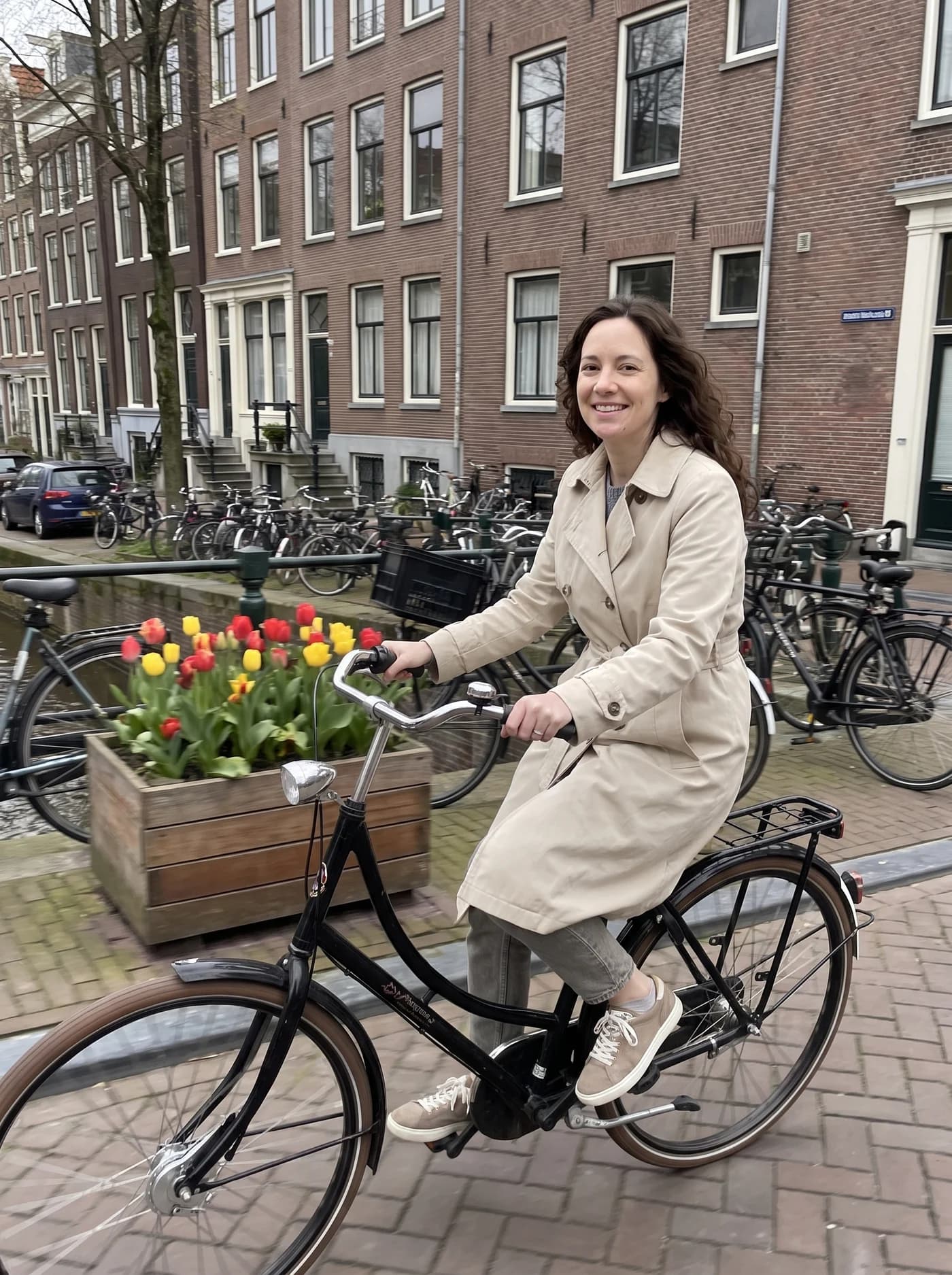 The same woman in a beige trench coat, cycling along an Amsterdam canal.